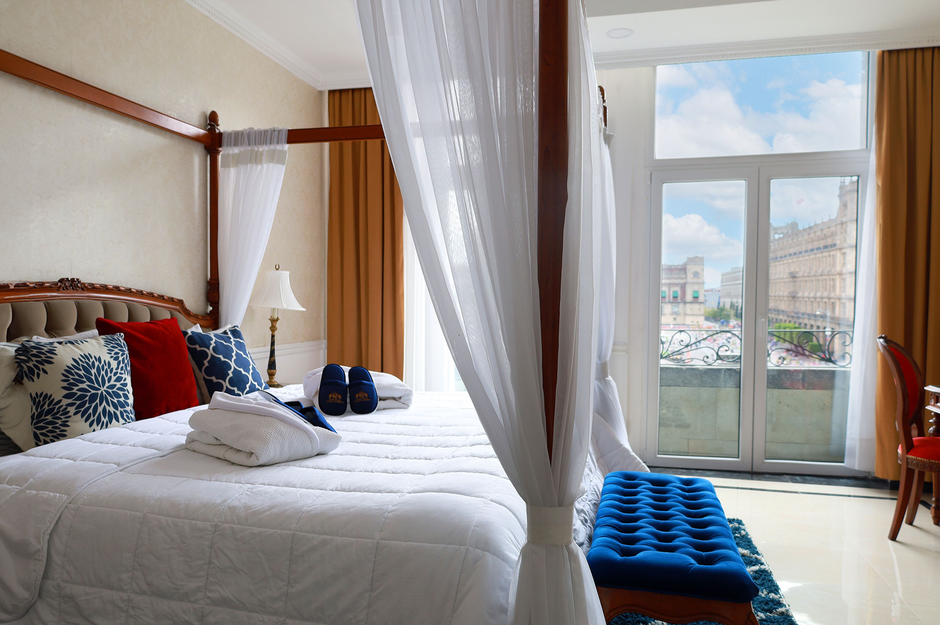 Ein Hotelzimmer mit einem großen Bett, einem Baldachin, Blick auf eine Stadtlandschaft durch einen Balkon und einem Sessel am Fenster.