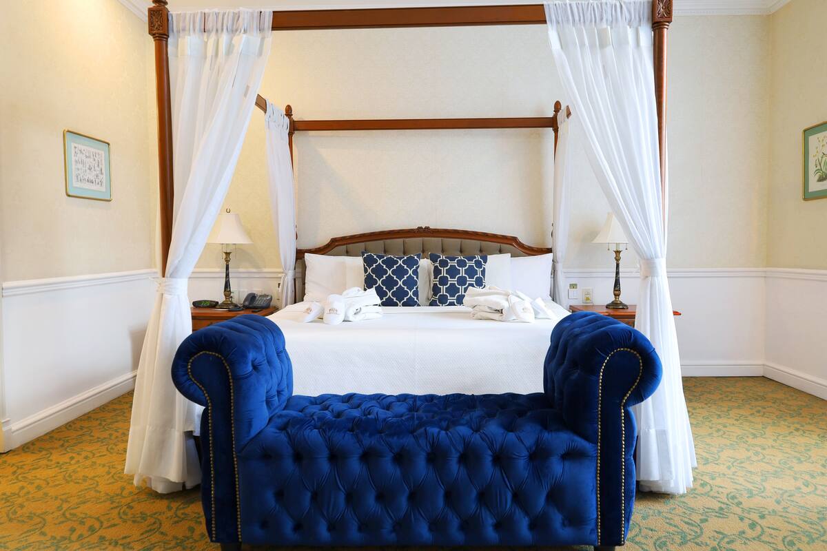 A four-poster canopy bed with white bedding, two bedside tables with lamps, and a blue upholstered bench.