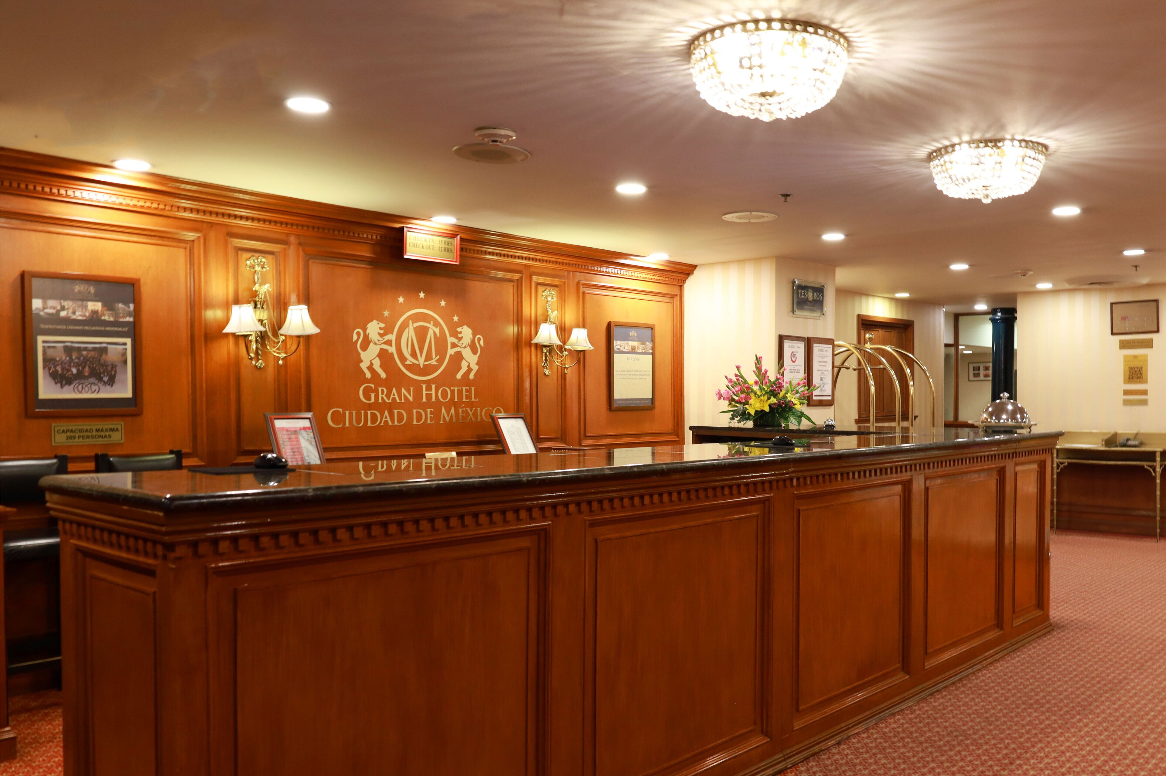 A reception desk at the Gran Hotel Ciudad de México with a wooden finish, chandeliers, and framed pictures on the walls.