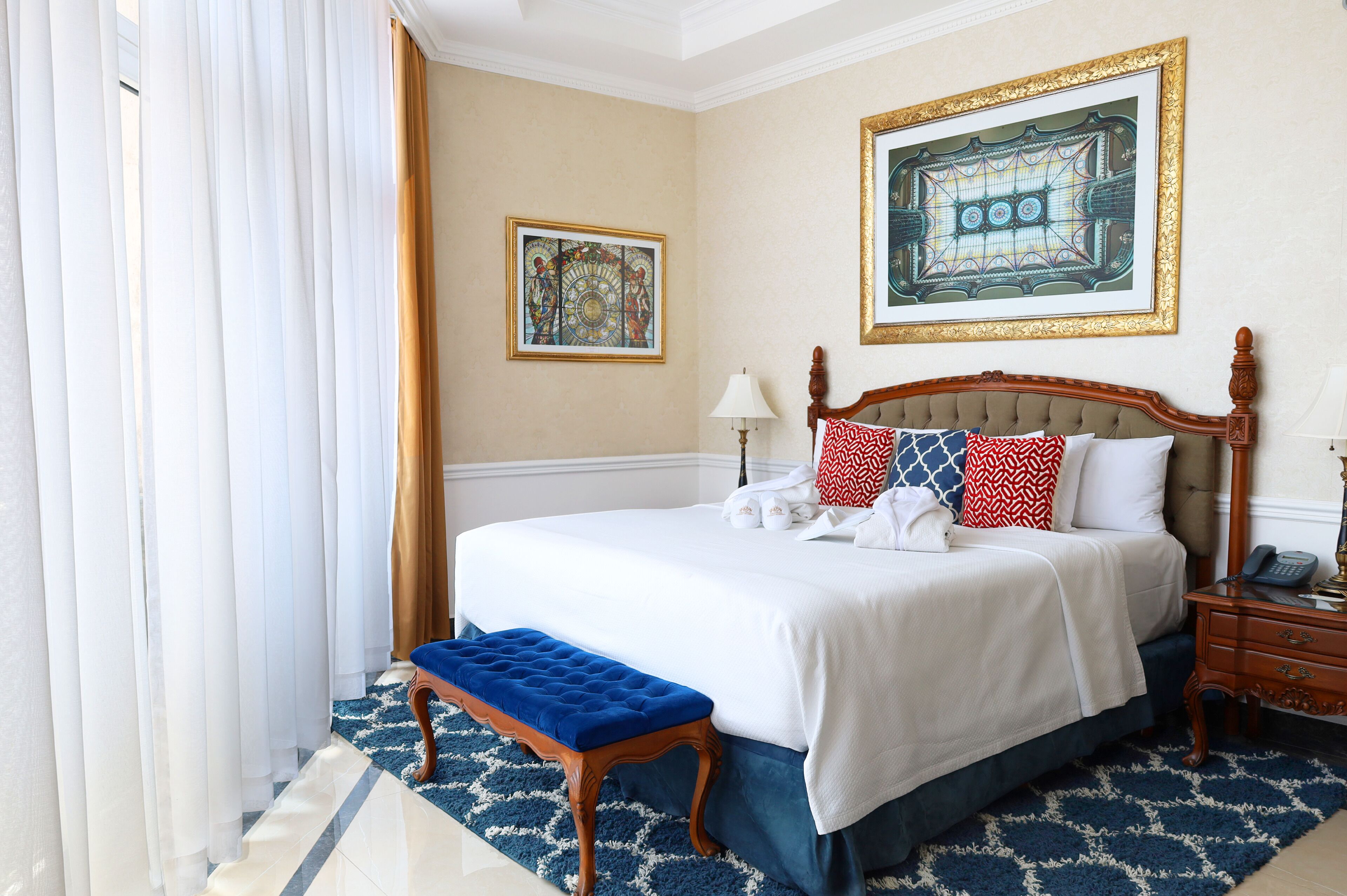 Habitación de hotel con una cama grande, una mesita de noche de madera, un banco azul y dos cuadros enmarcados en la pared.