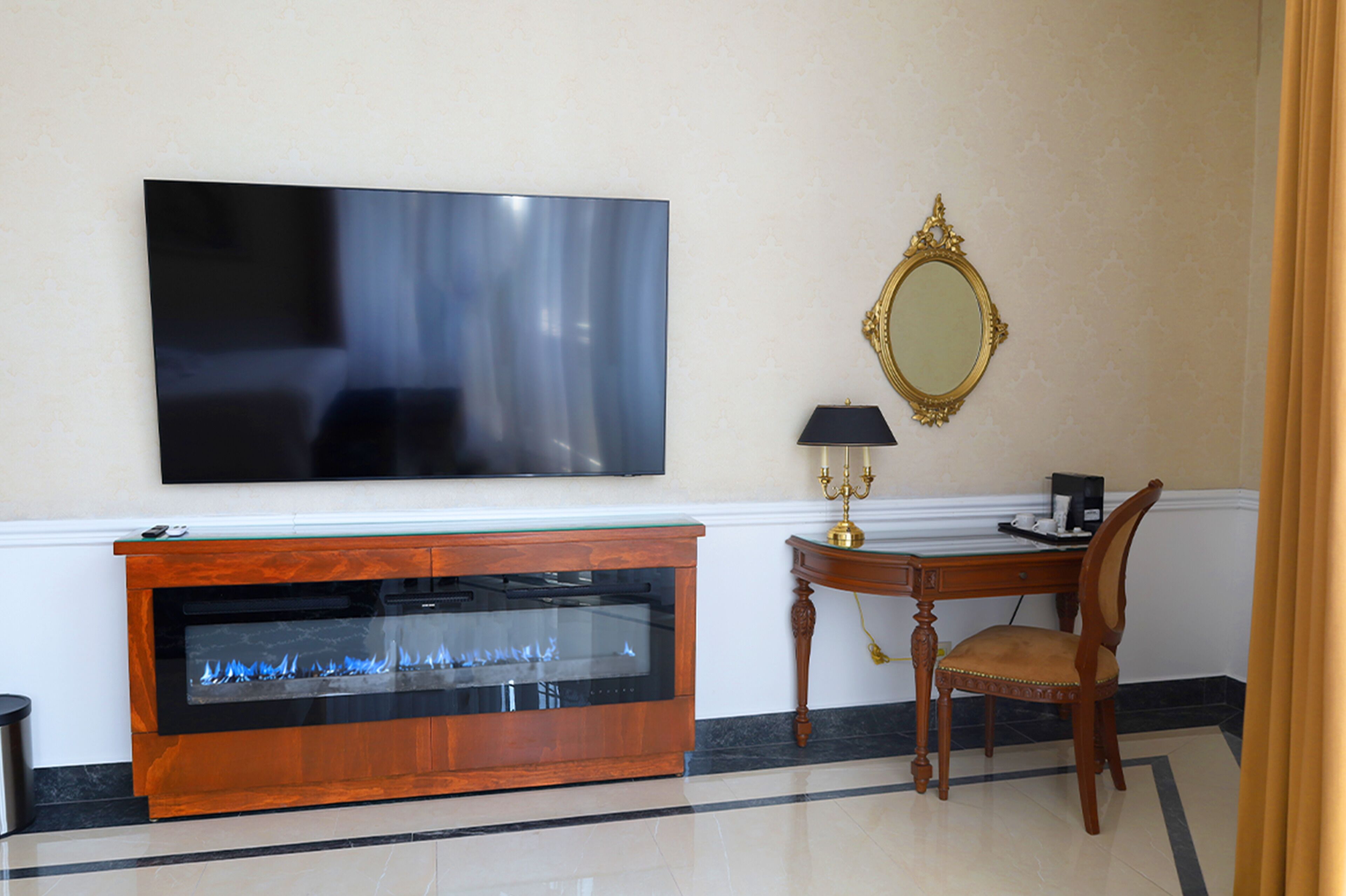A modern living room with a flat-screen TV, a glass fireplace, a wooden sideboard, a table with a lamp, and a mirror on the wall.