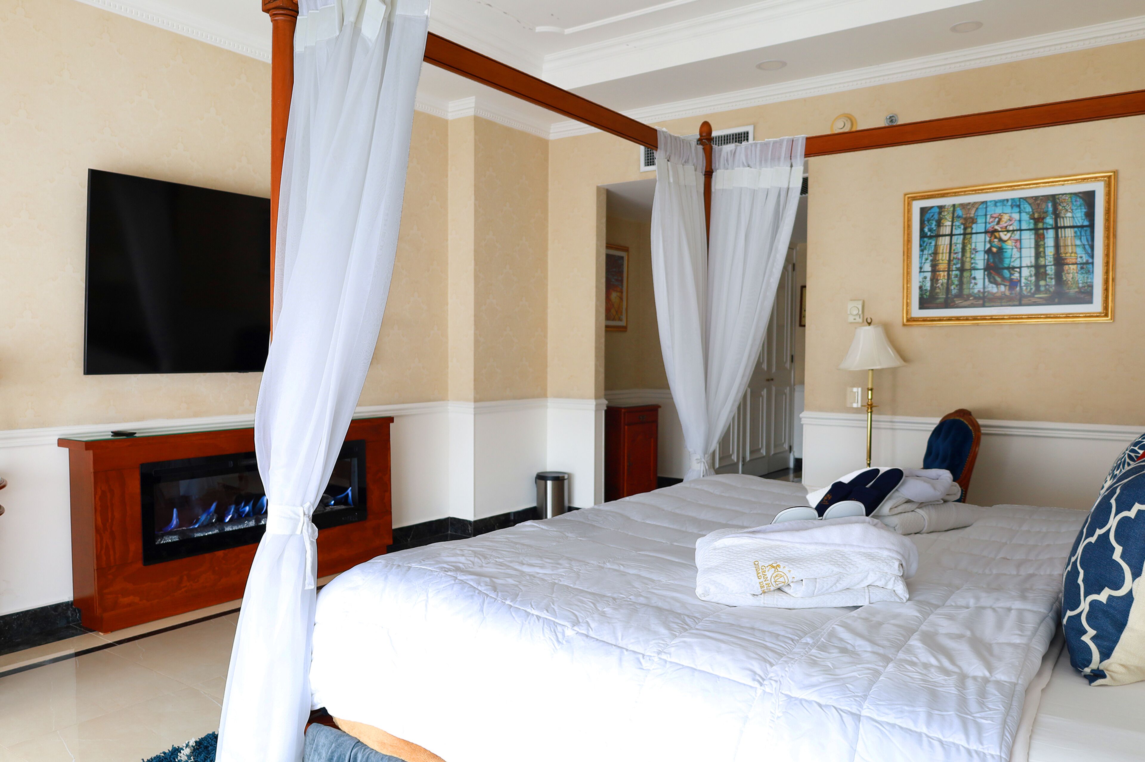 A hotel room with a large bed, a flat-screen TV mounted on the wall, a fireplace, and a painting on the wall.