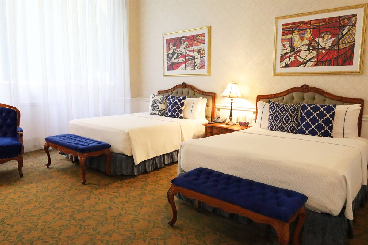 A hotel room with two beds, a chair, and a bench. There are two paintings on the wall and a lamp on the nightstand.