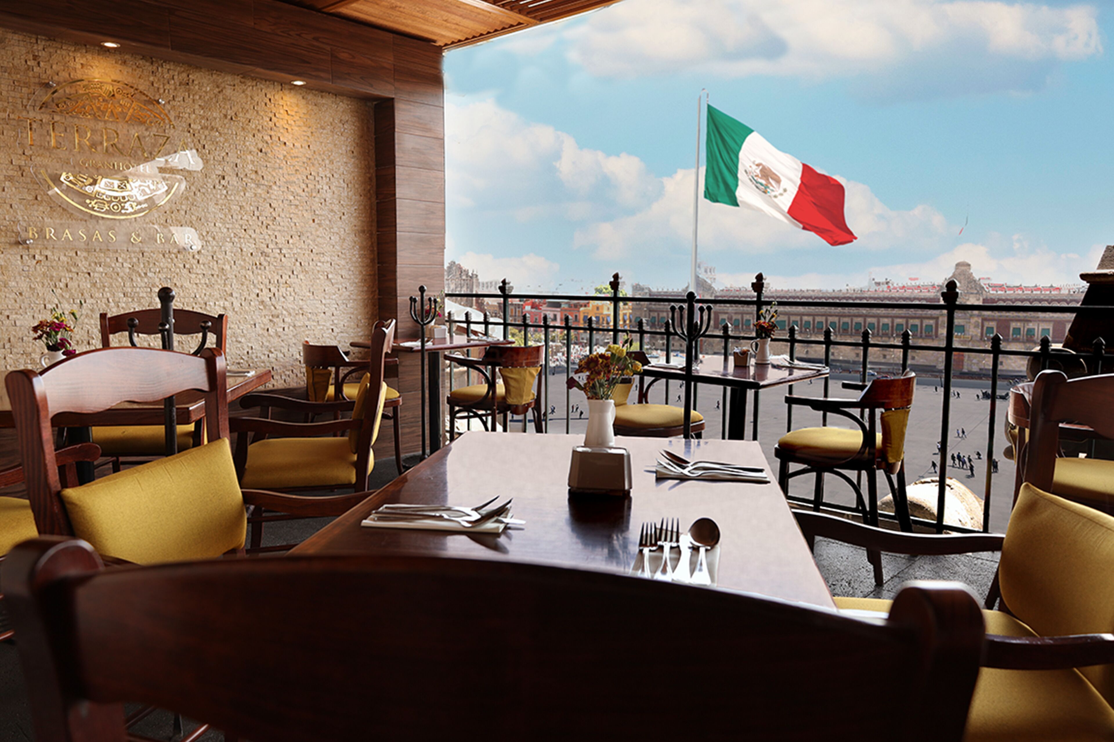 Un restaurante con terraza que da a un cuerpo de agua, con una bandera y vista a un paisaje urbano lejano.