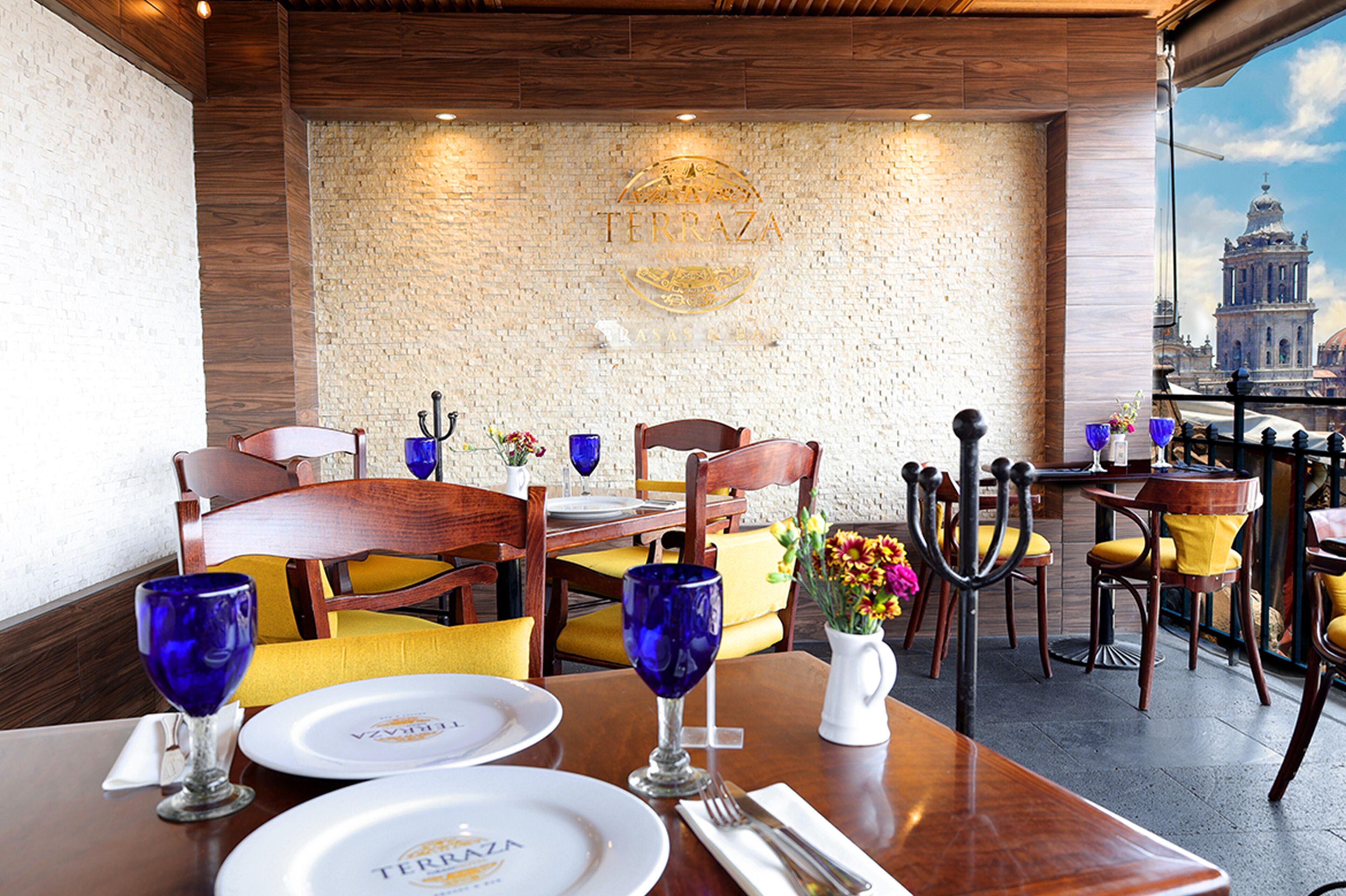 Un restaurante con mesas y sillas de madera, una pared de piedra con un logotipo y vista a un edificio a través de una ventana.