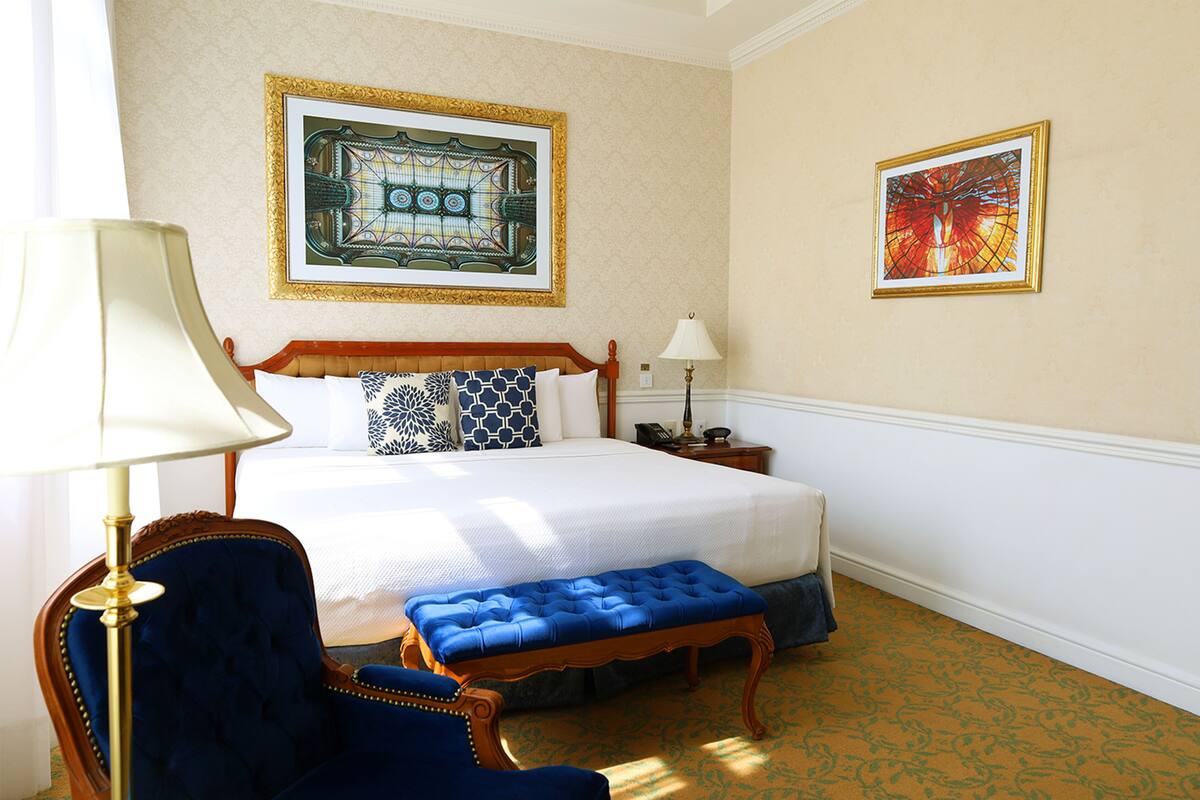 A hotel room with a large bed, a blue upholstered chair, a wooden bench, two bedside tables, and two wall-mounted lamps.