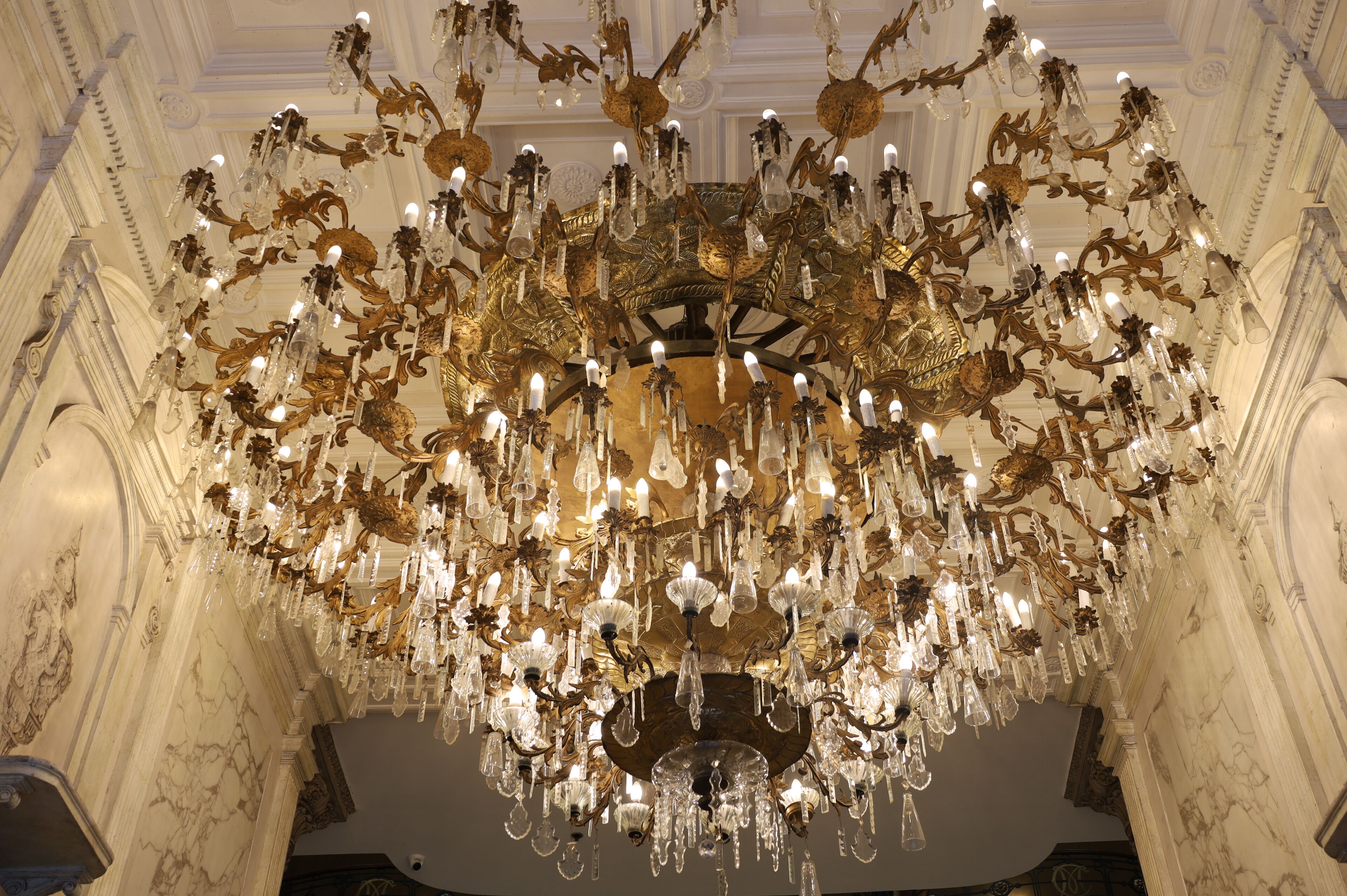 Un gran candelabro con varios niveles y detalles de cristal en una amplia sala con arquitectura clásica.