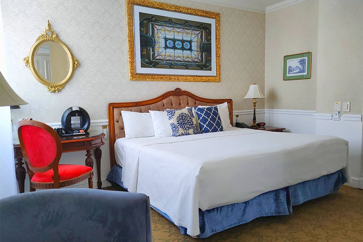 A hotel room with a large bed, a wooden headboard, a red chair, a desk with a lamp, and a framed artwork on the wall.