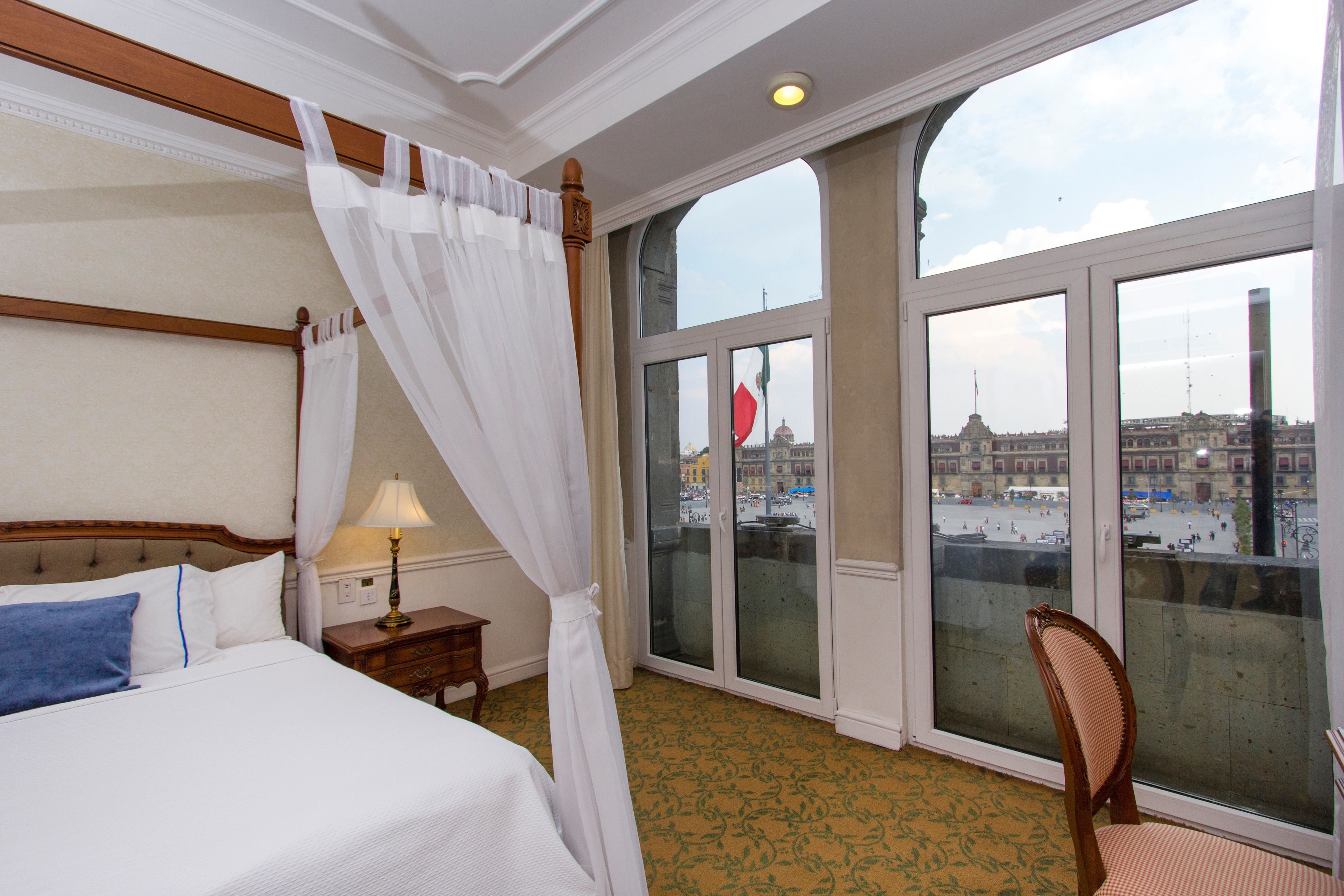 A hotel room with a large bed, a chair, a nightstand, and a view of a cityscape through large windows.
