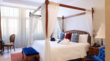 Un lit à baldaquin à quatre poteaux avec du linge de lit blanc, un coussin aux motifs rouges et bleus, une table de chevet en bois avec une lampe et un fauteuil bleu.