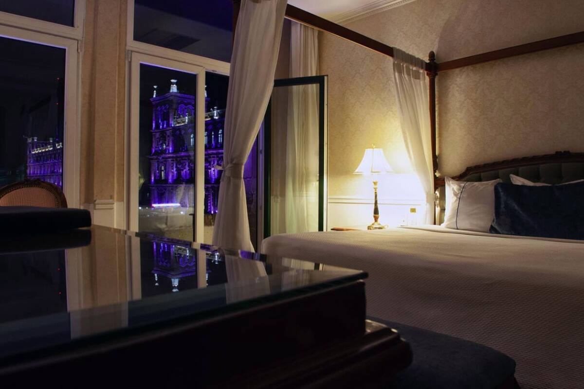A hotel room with a bed, a nightstand, a lamp, and a view of a lit-up building outside.