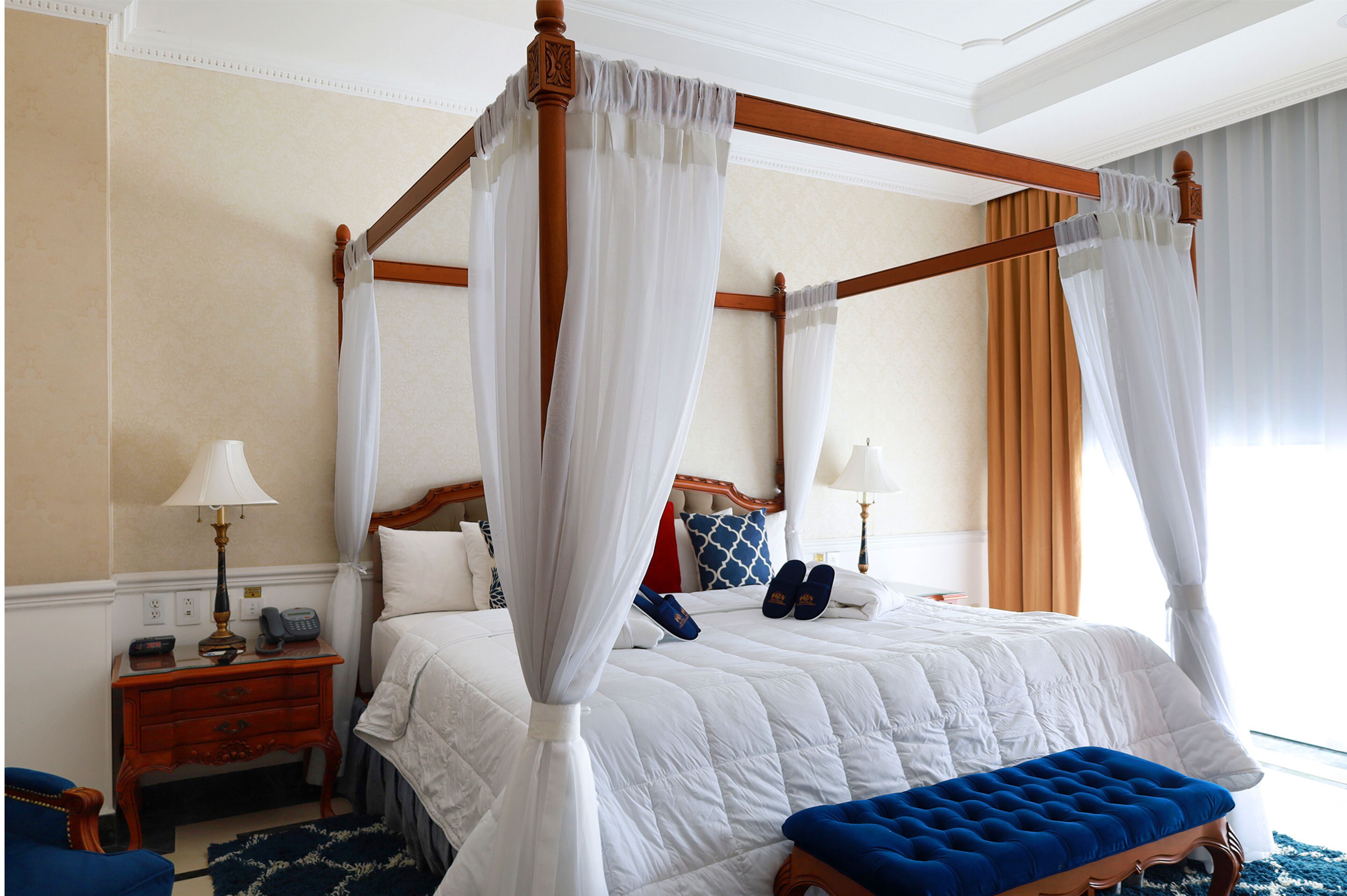 Una cama con dosel y cuatro columnas, ropa de cama blanca, un banco de madera y una mesita de noche con lámpara y teléfono.