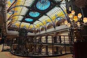 Un interior ornamentado con techo de vitrales, barandillas de hierro forjado y una araña.