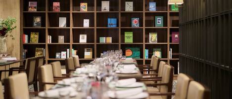Un restaurante con una larga mesa de comedor preparada para una comida, rodeada de sillas, y una estantería llena de libros al fondo.