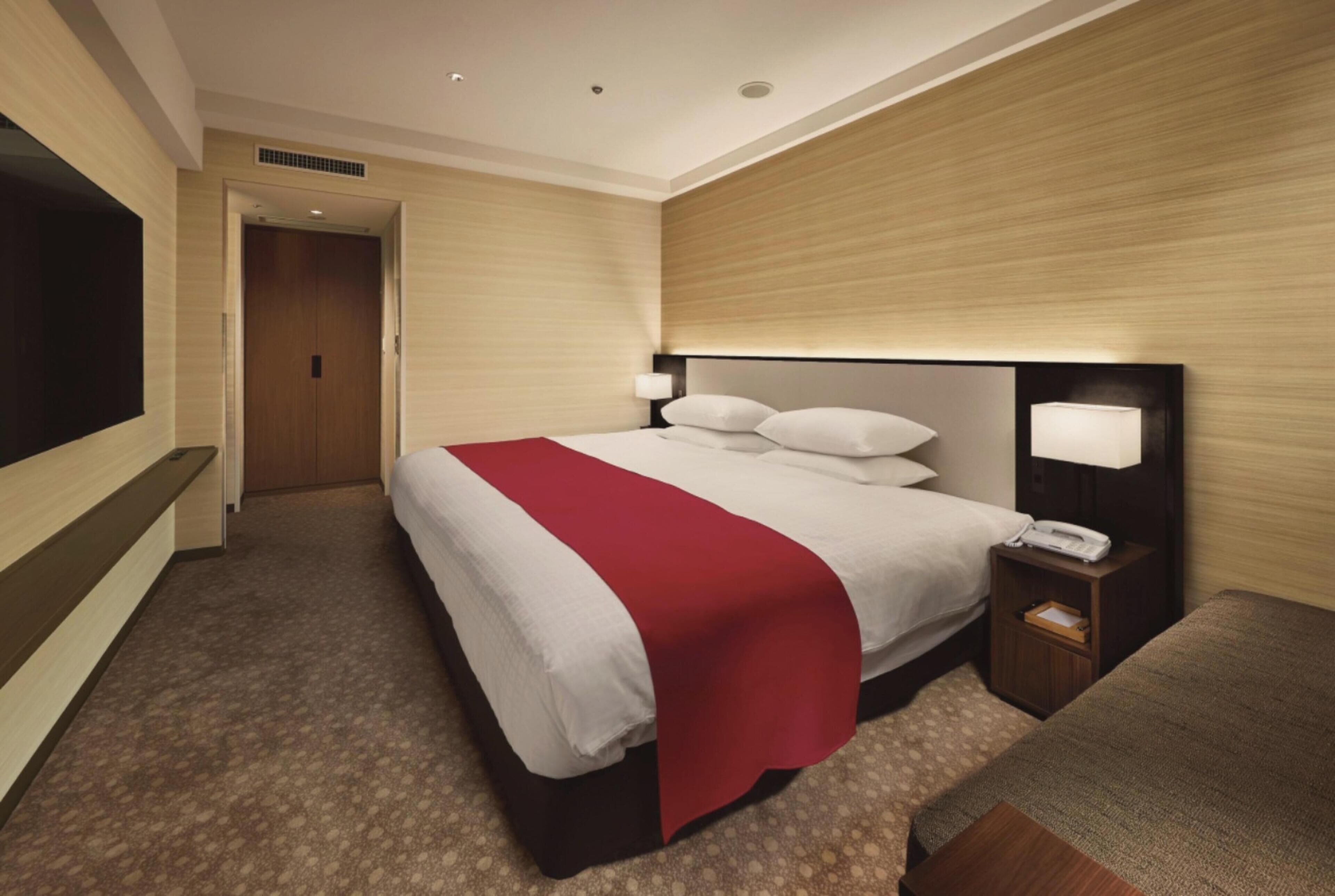 A hotel room with a large bed, a bedside table with a lamp, a TV mounted on the wall, and a door leading to another room.