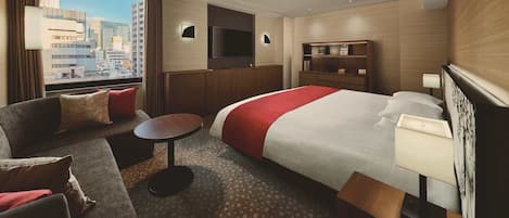 Habitación de hotel con una cama grande, un sofá, una mesita, televisión y vista a la ciudad.