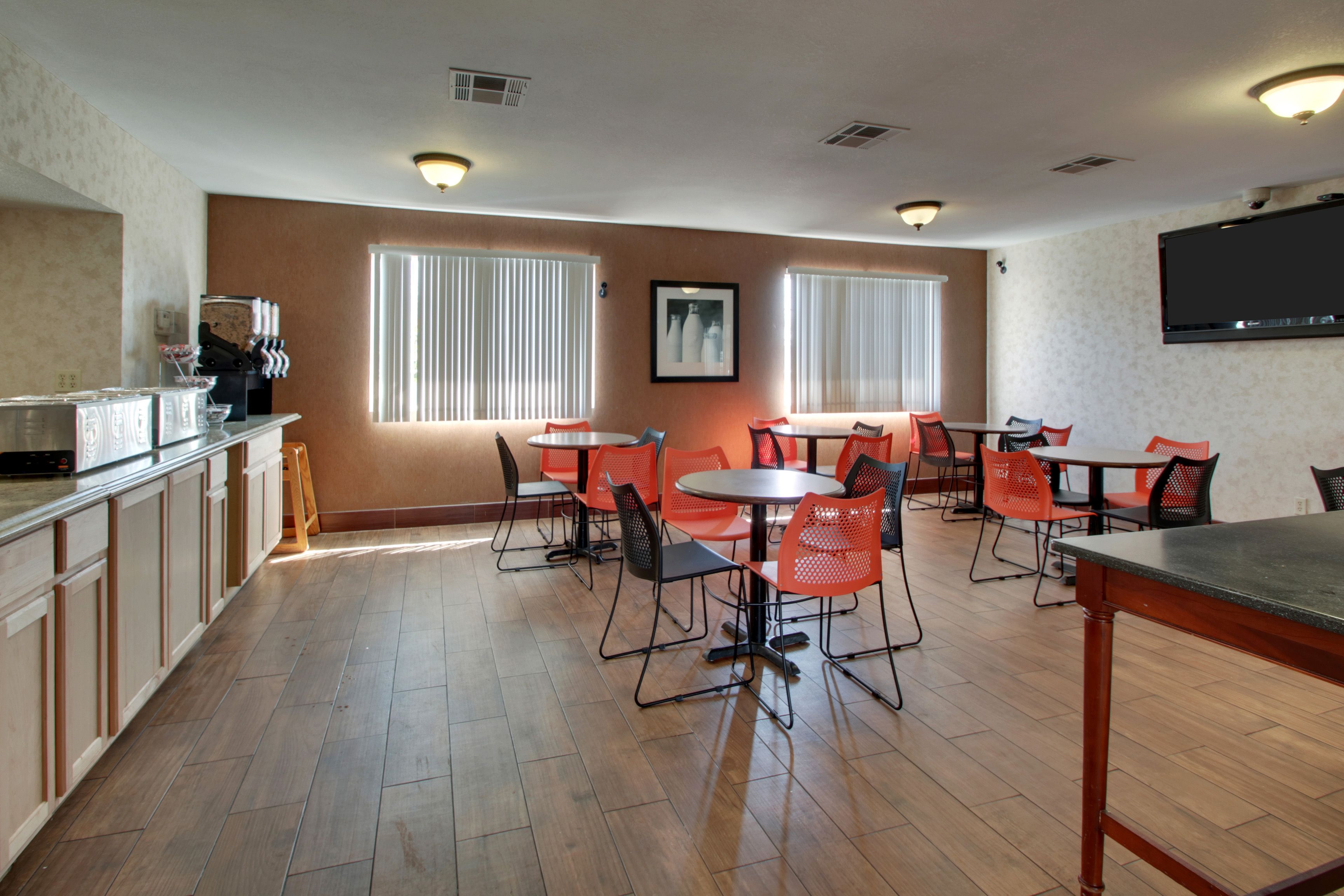 A dining area with multiple tables and chairs, a kitchen area with cabinets and appliances, and large windows with blinds.
