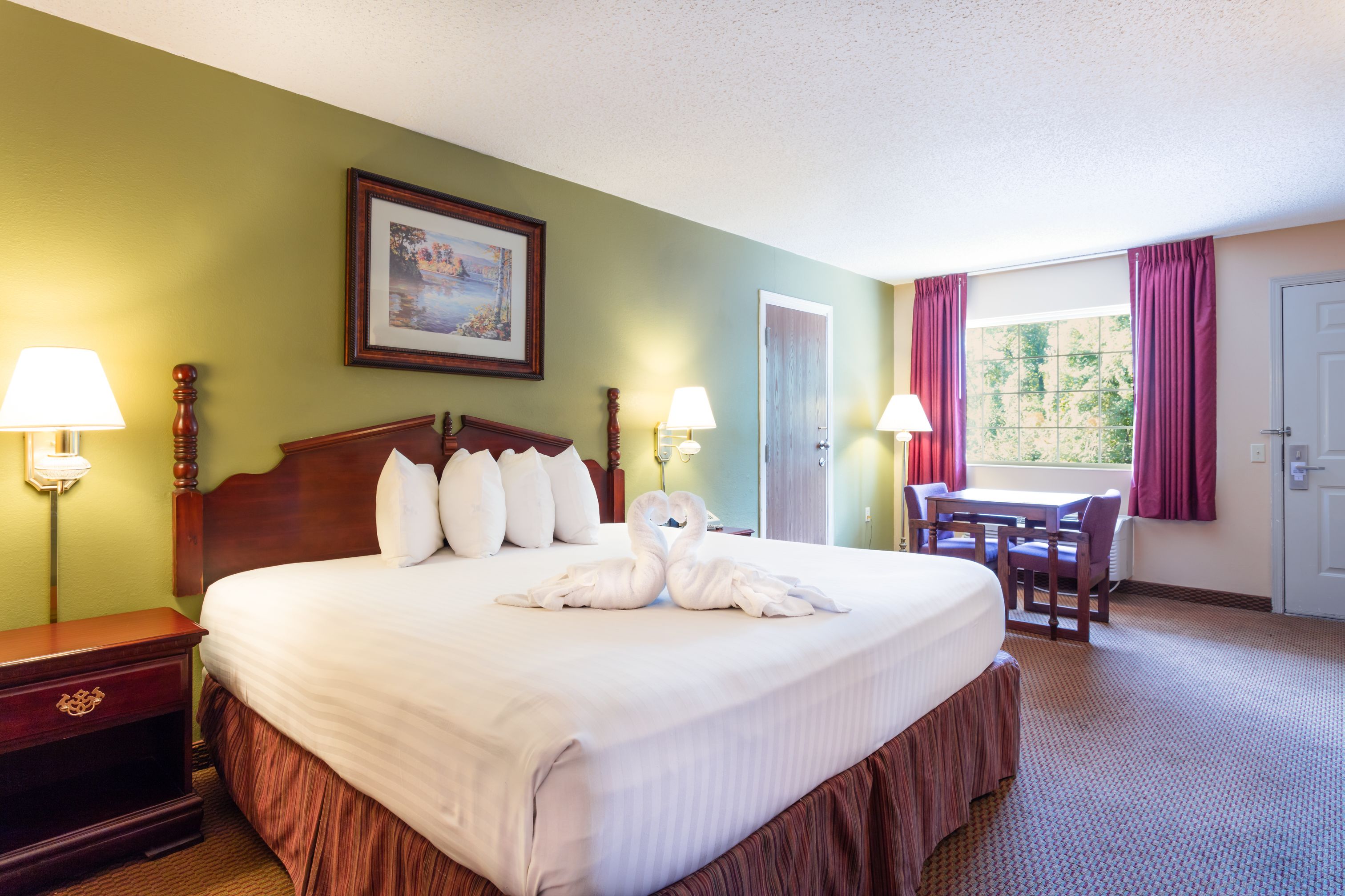 A hotel room with a large bed, two bedside lamps, a wooden headboard, a framed picture on the wall, a dining table with chairs, and a window with curtains.