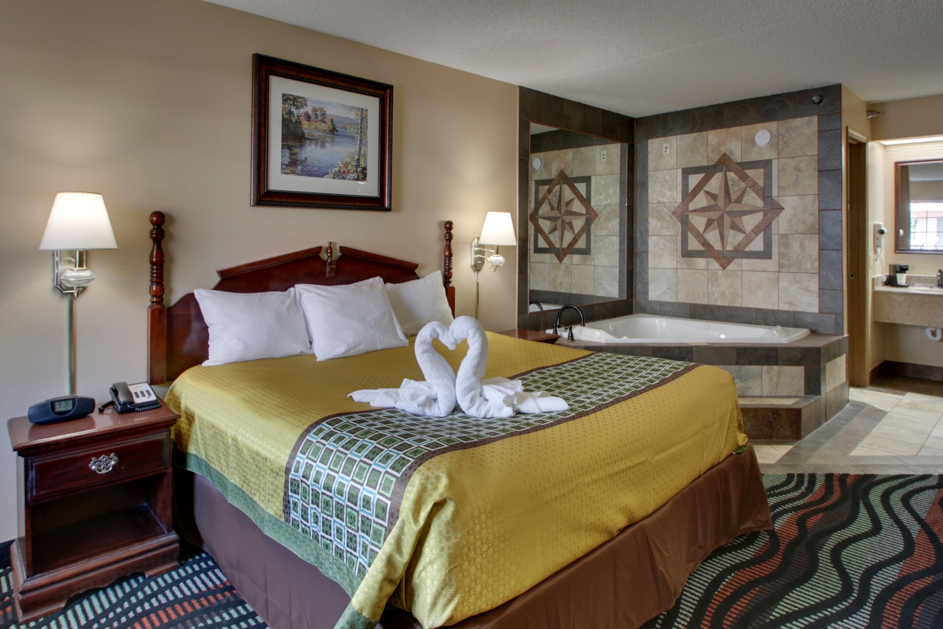 A hotel room with a large bed, two bedside lamps, a framed picture on the wall, a bathtub, and a bathroom with a large mirror.