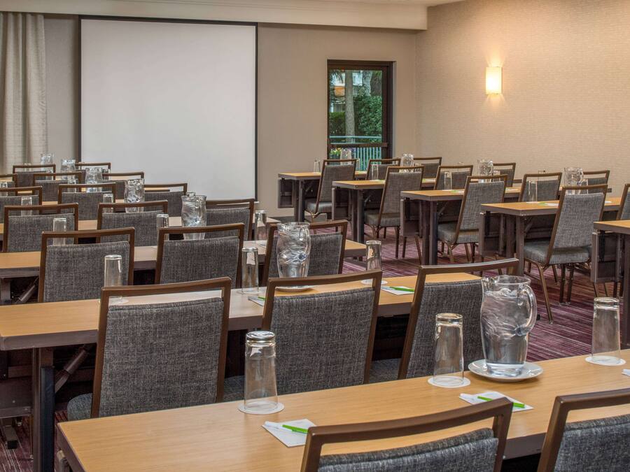 Un salón de conferencias con varias mesas y sillas, una pantalla de proyector y un ventanal con vistas a árboles.
