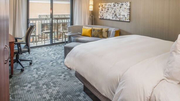 A hotel room with a large bed, a desk, a chair, a balcony with a table and chairs, and a wall-mounted lamp. - Sonesta Select Seattle Bellevue Redmond (Bellevue)