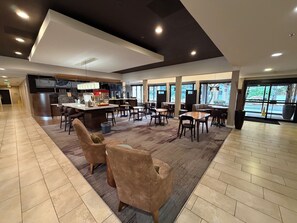 Un amplio vestíbulo de hotel con una zona de bar central, arreglos de asientos y ventanas grandes que ofrecen luz natural.