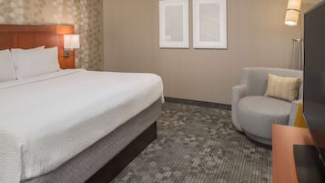 Une chambre d’hôtel avec un grand lit, un fauteuil gris, un téléviseur à écran plat et deux tableaux encadrés au mur.