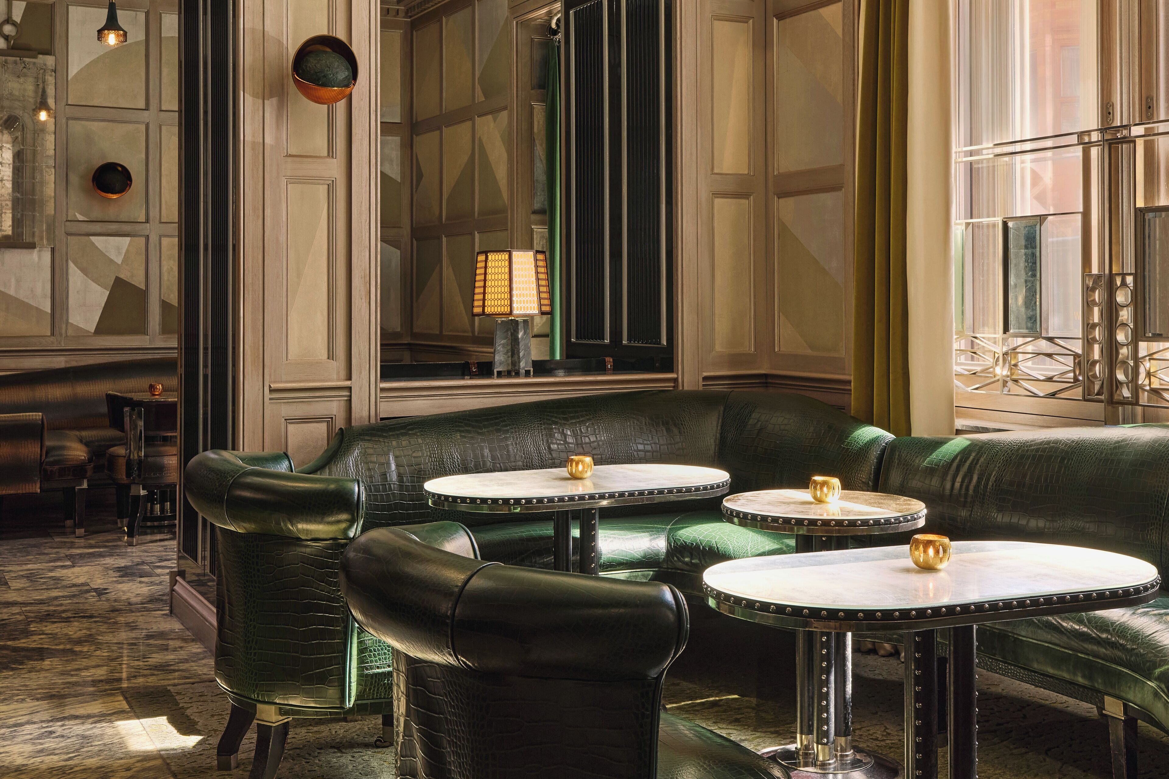 A green leather sofa and two round tables with gold accents in a well-lit room with a patterned floor and a large mirror.