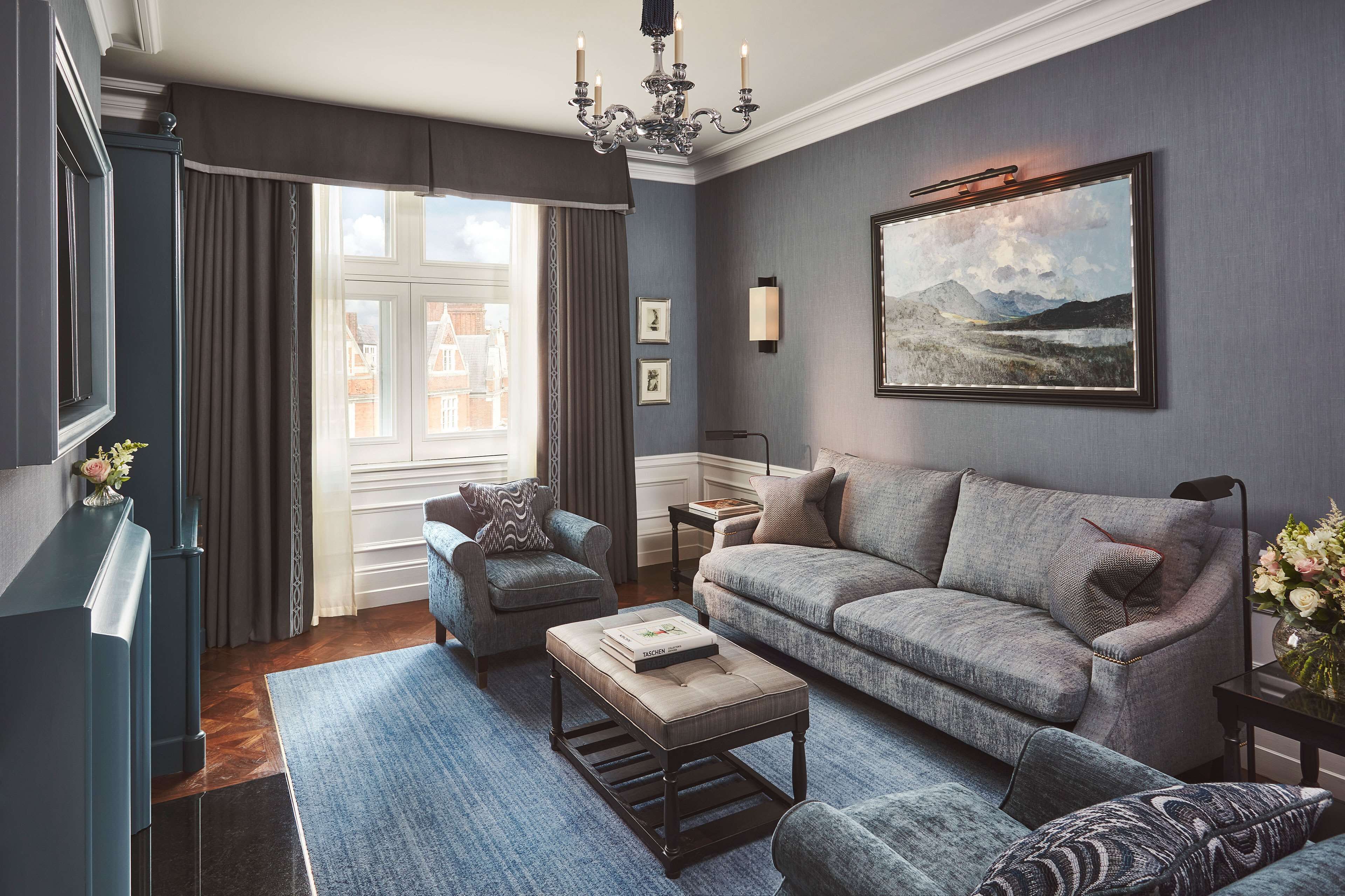 A modern living room with a grey sofa, a blue armchair, and a coffee table. There is a painting on the wall and a window with curtains.