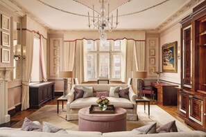 A spacious living room with a large sofa, a coffee table with flowers, a chandelier, and a view of buildings through large windows.
