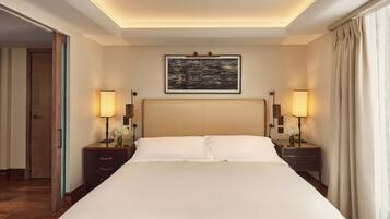 Een hotelkamer met een groot bed, twee nachtkastjes met lampen, een lijst met kunstwerk boven het bed, en een deur die naar een andere kamer leidt.