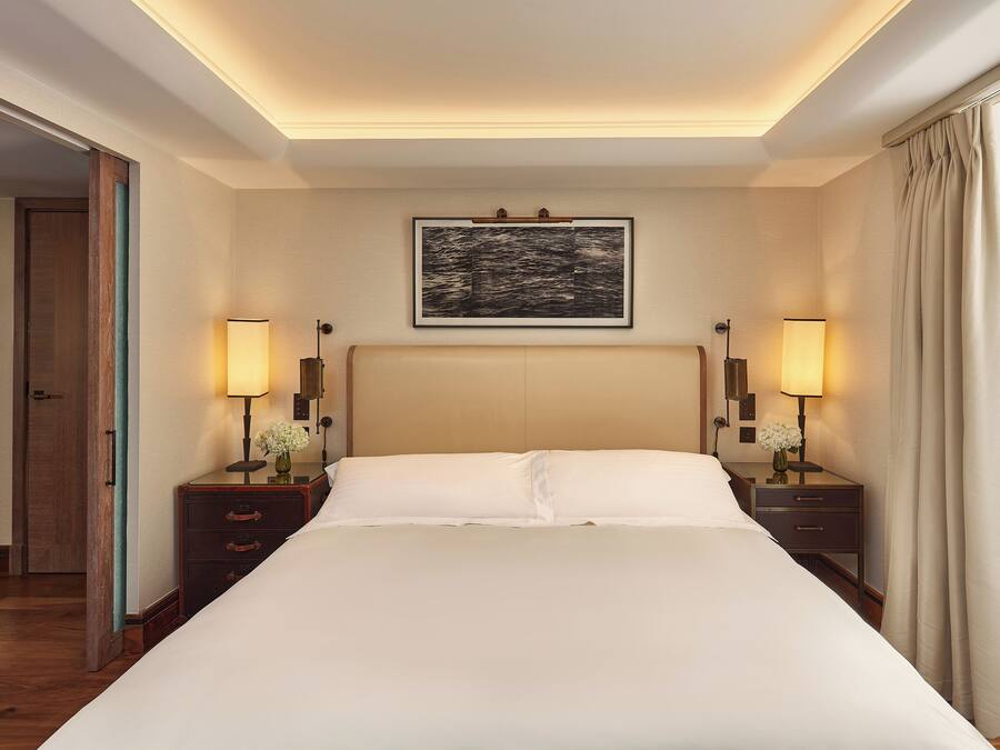 Een hotelkamer met een groot bed, twee nachtkastjes met lampen, een lijst met kunstwerk boven het bed, en een deur die naar een andere kamer leidt.