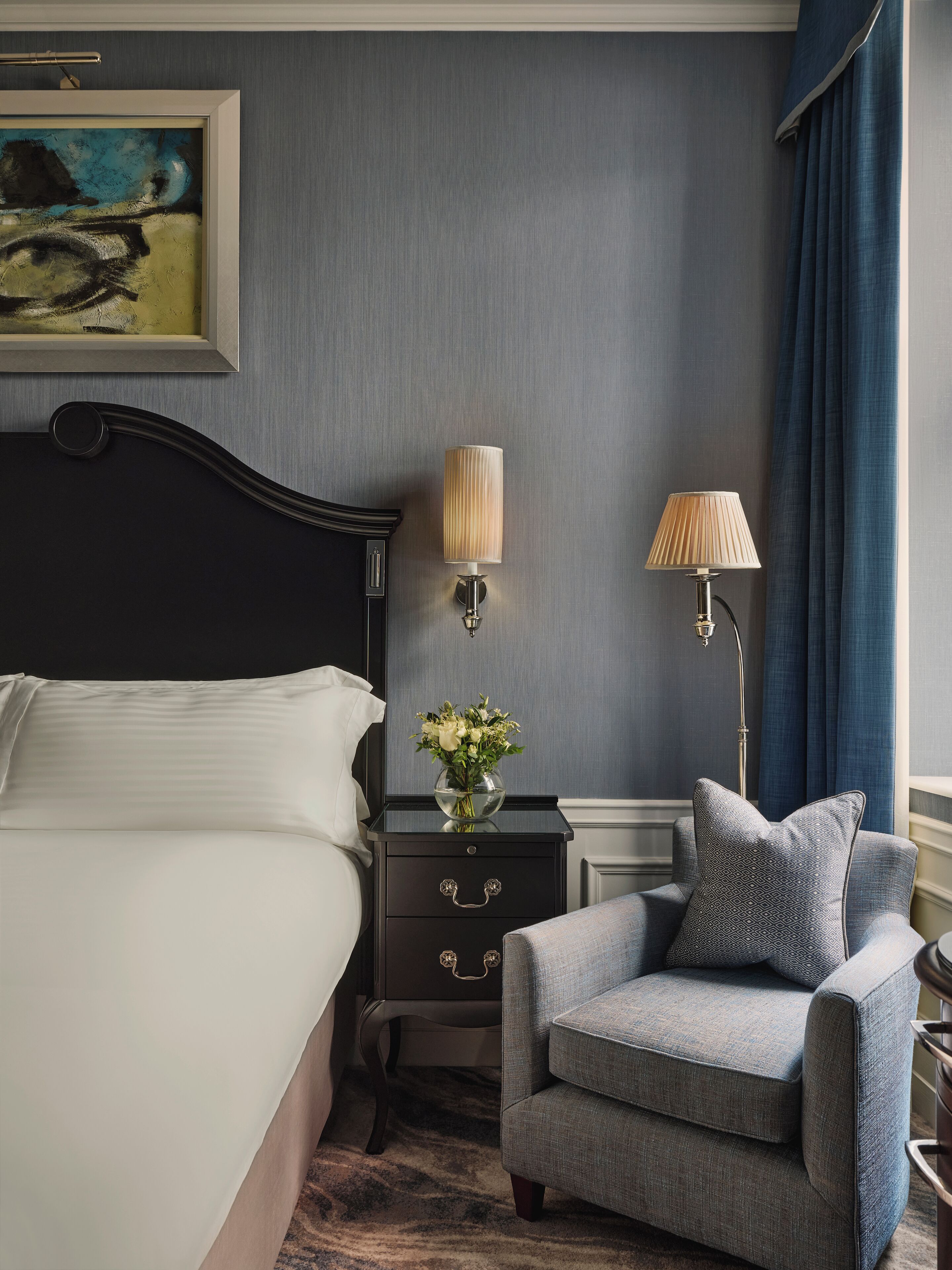 A neatly arranged bedroom with a bed, a nightstand with a lamp, a chair, and a vase of flowers.