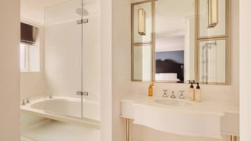 Un bany modern amb plat de dutxa envoltat de vidre, un lavabo blanc amb detalls daurats i un mirall gran amb adorns daurats.