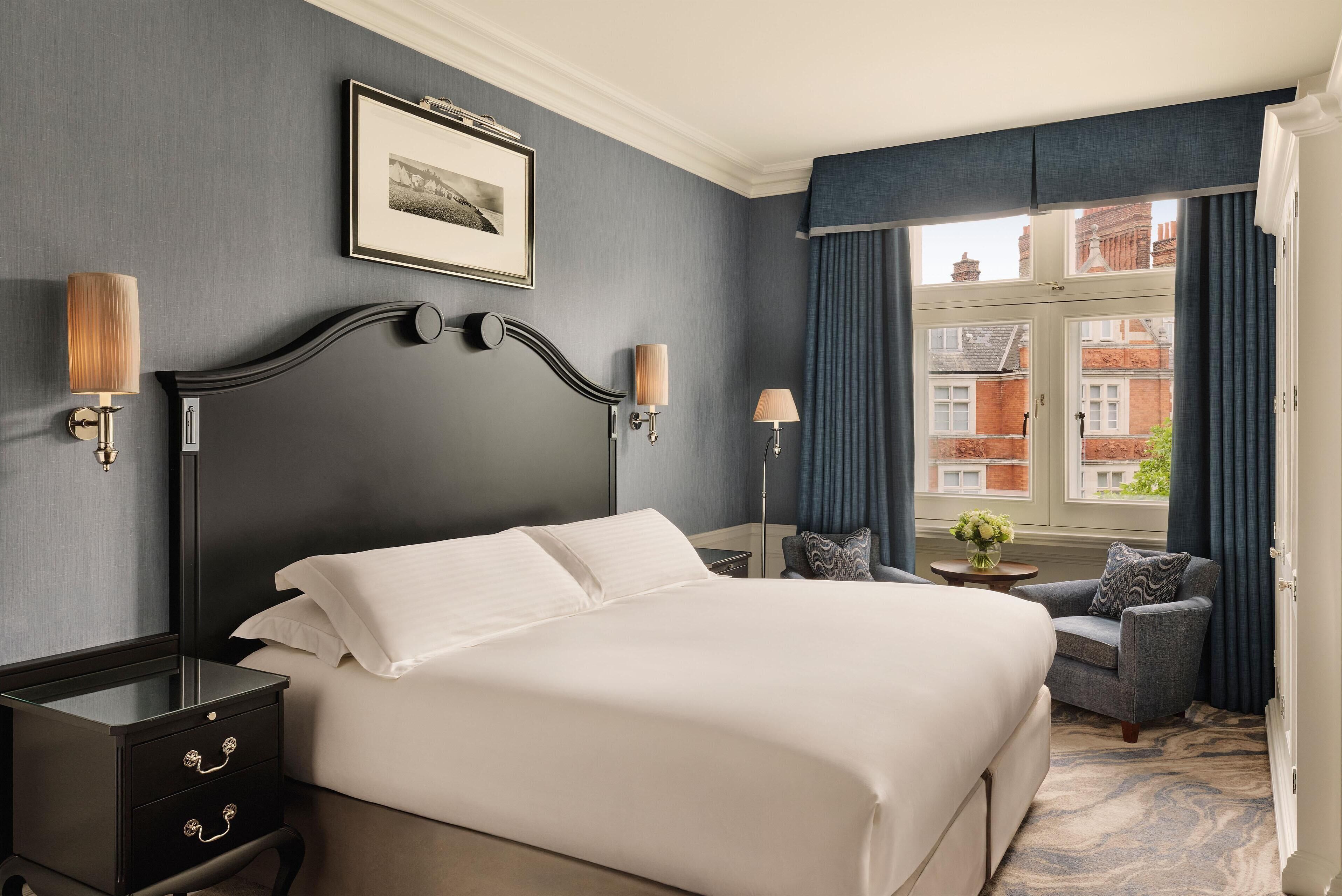 Quarto de hotel com uma cama grande, duas luminárias de cabeceira, um quadro na parede, uma cadeira e vista para edifícios pela janela.