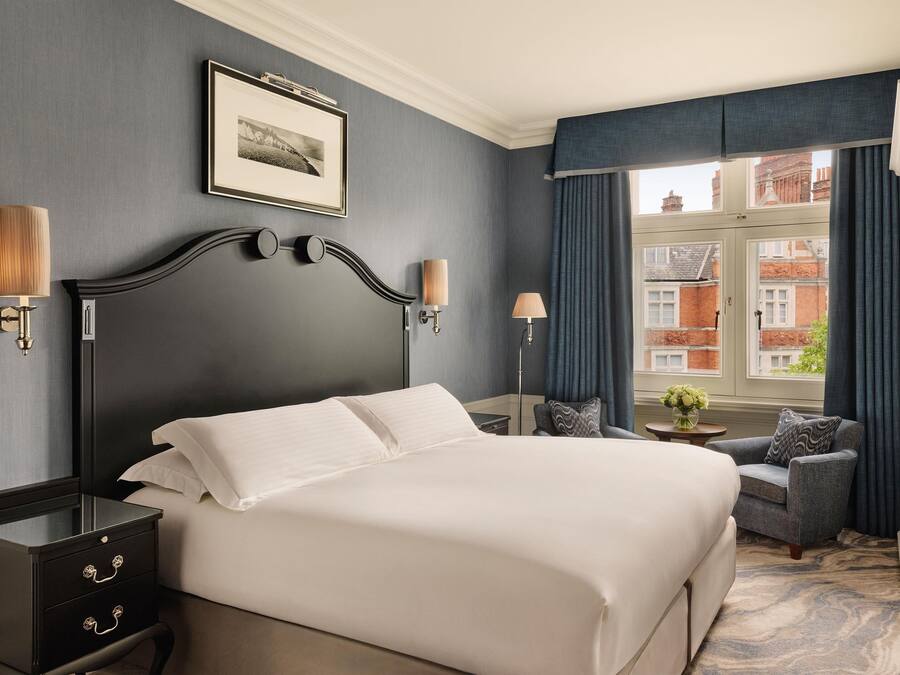 Quarto de hotel com uma cama grande, duas luminárias de cabeceira, um quadro na parede, uma cadeira e vista para edifícios pela janela.