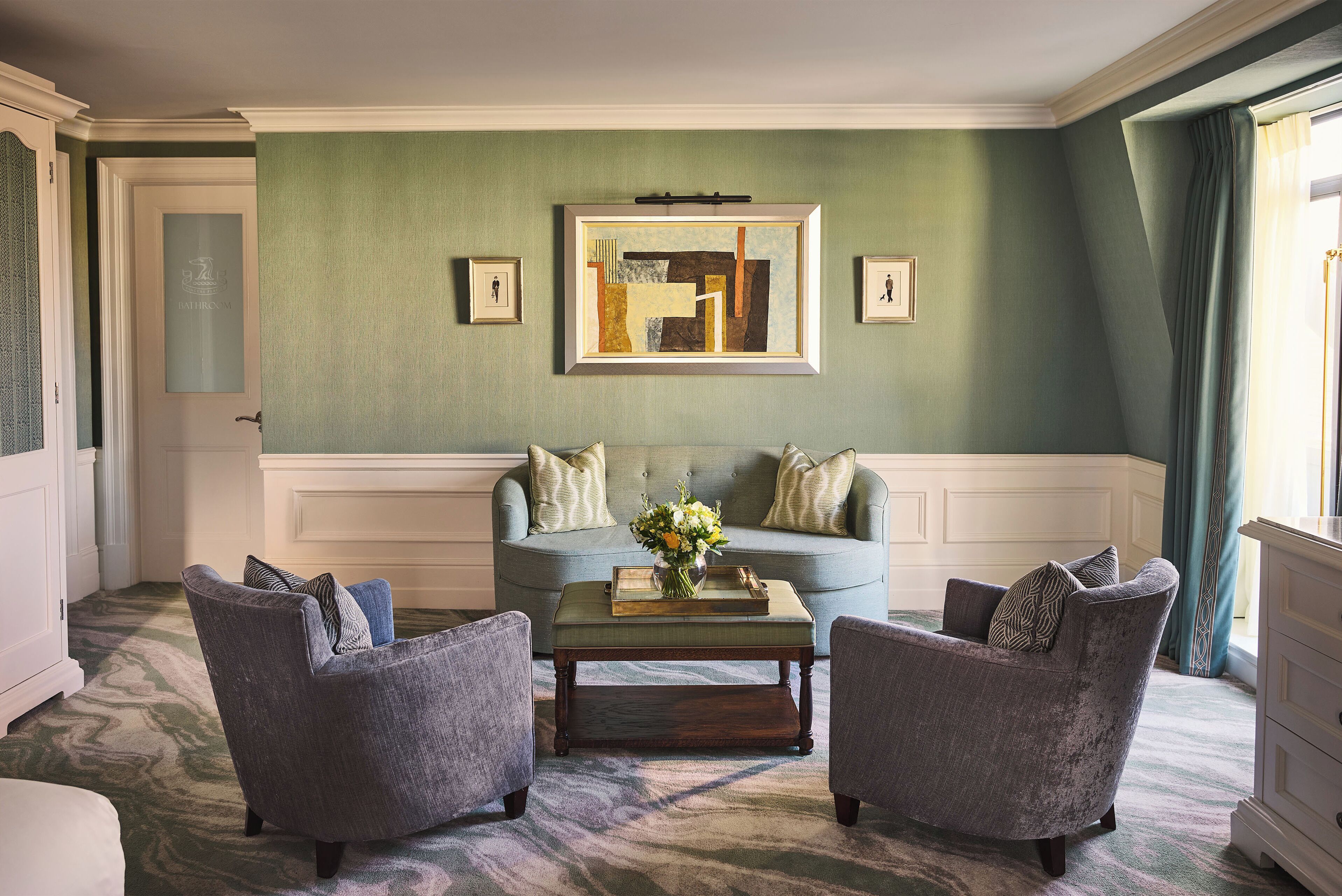 A living room with a green wall, a framed abstract painting, a sofa with pillows, two armchairs, and a coffee table with a vase of flowers.
