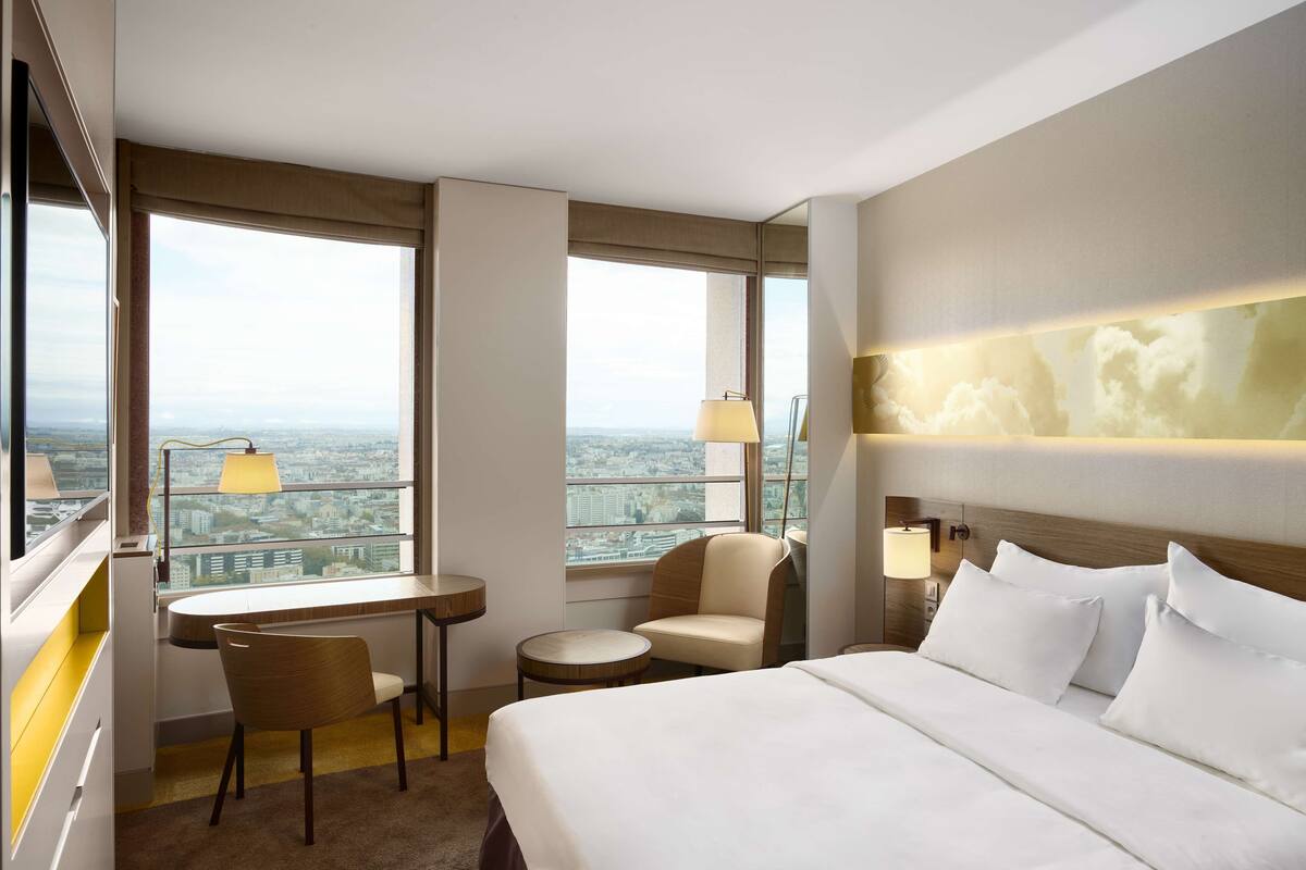 A hotel room with a bed, a desk, a chair, a small table, and a view of the cityscape.