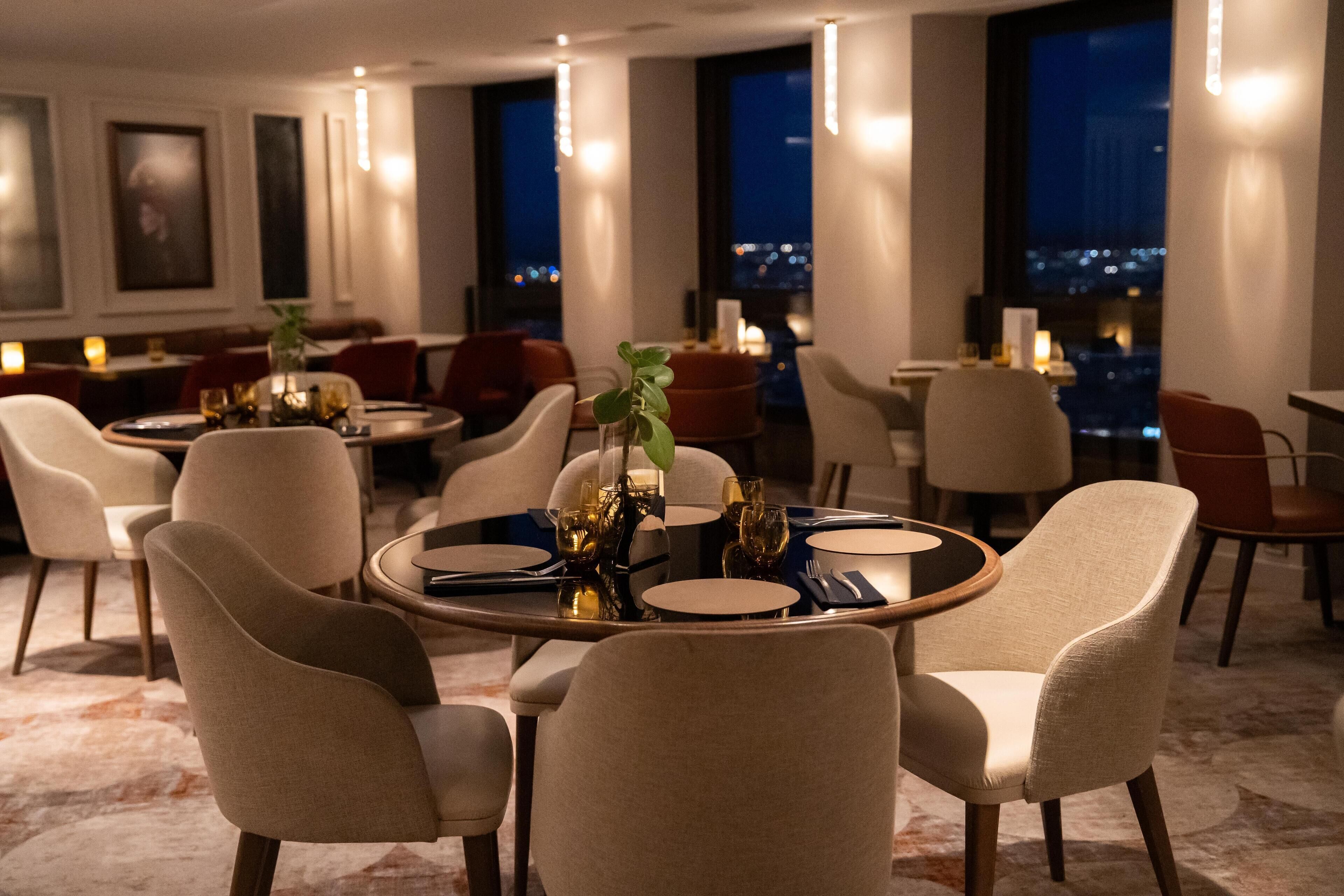 A modern dining area with round tables, beige chairs, and a city view through large windows.