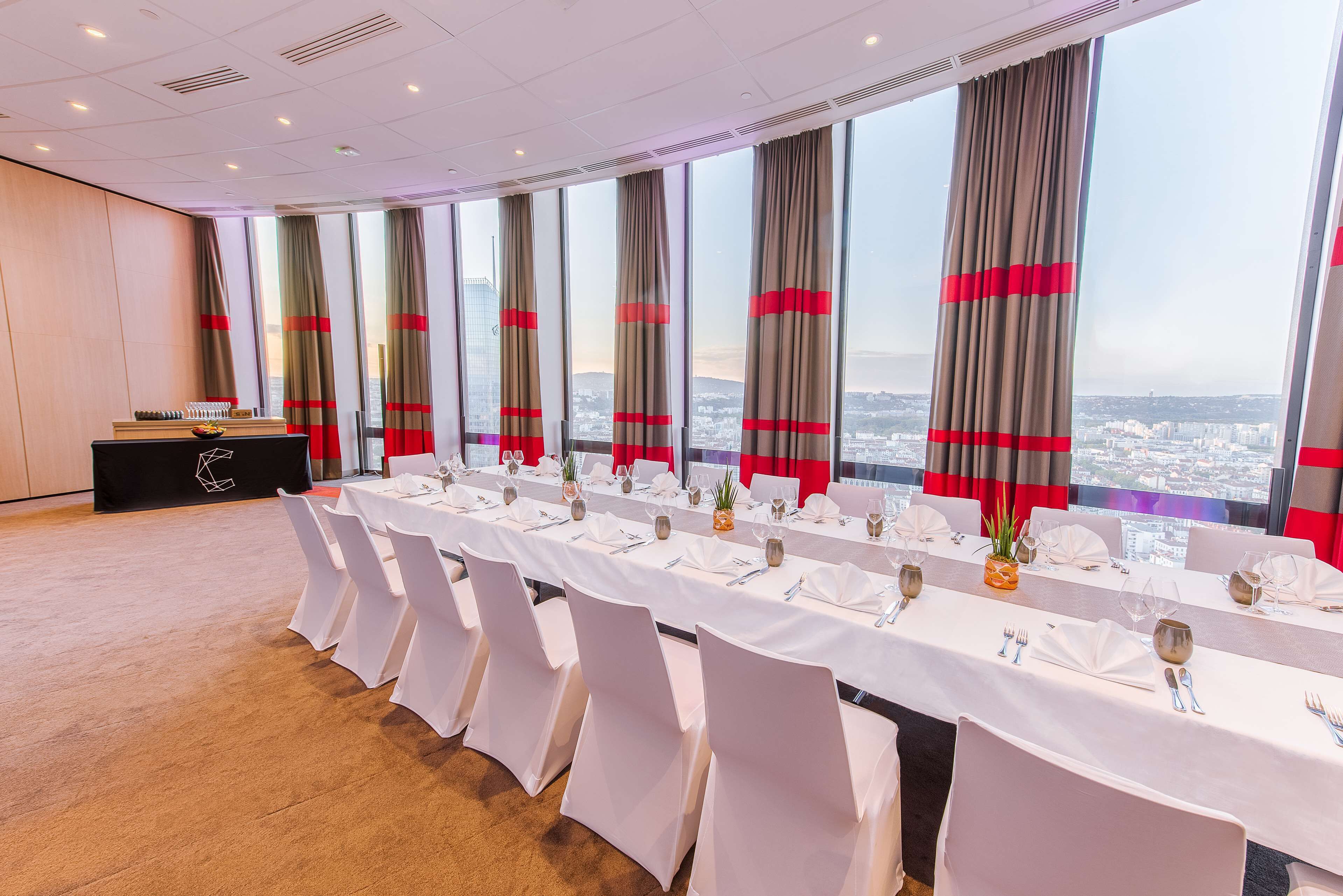 A conference room with a long table set for a meeting, large windows with curtains, and a city view.