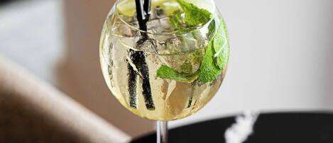 A cocktail glass with a stemmed base, filled with a clear liquid, ice cubes, and garnished with a mint leaf, on a black table with a white crack.
