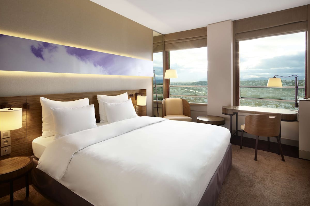 A hotel room with a large bed, two bedside tables, a desk, a chair, and a view of the city through large windows.