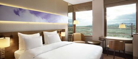 Una habitación de hotel con una cama grande, dos mesitas de noche, un escritorio, una silla y vistas de la ciudad a través de amplios ventanales.