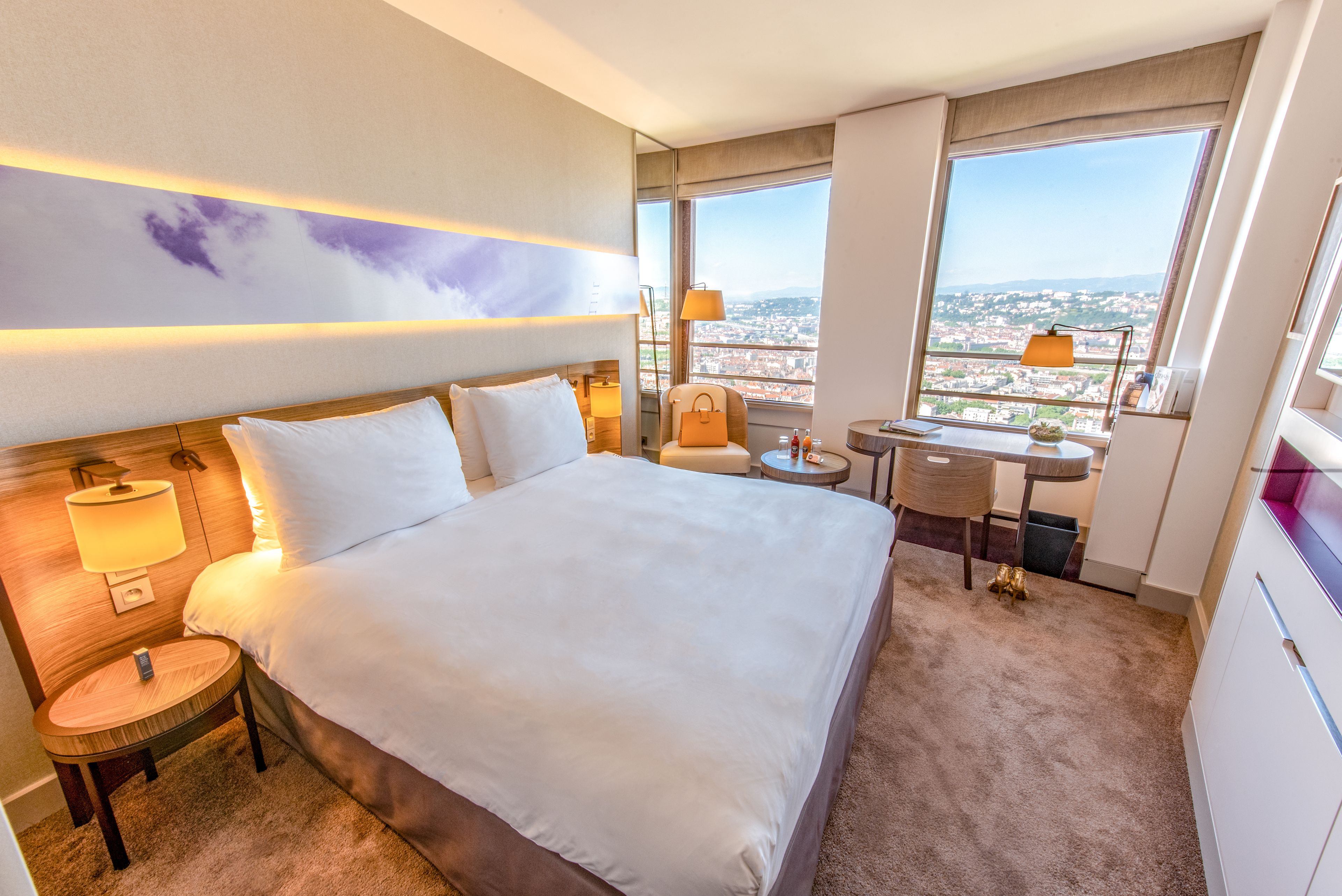 Habitación de hotel con una cama grande, un escritorio y vistas de la ciudad a través de amplias ventanas.