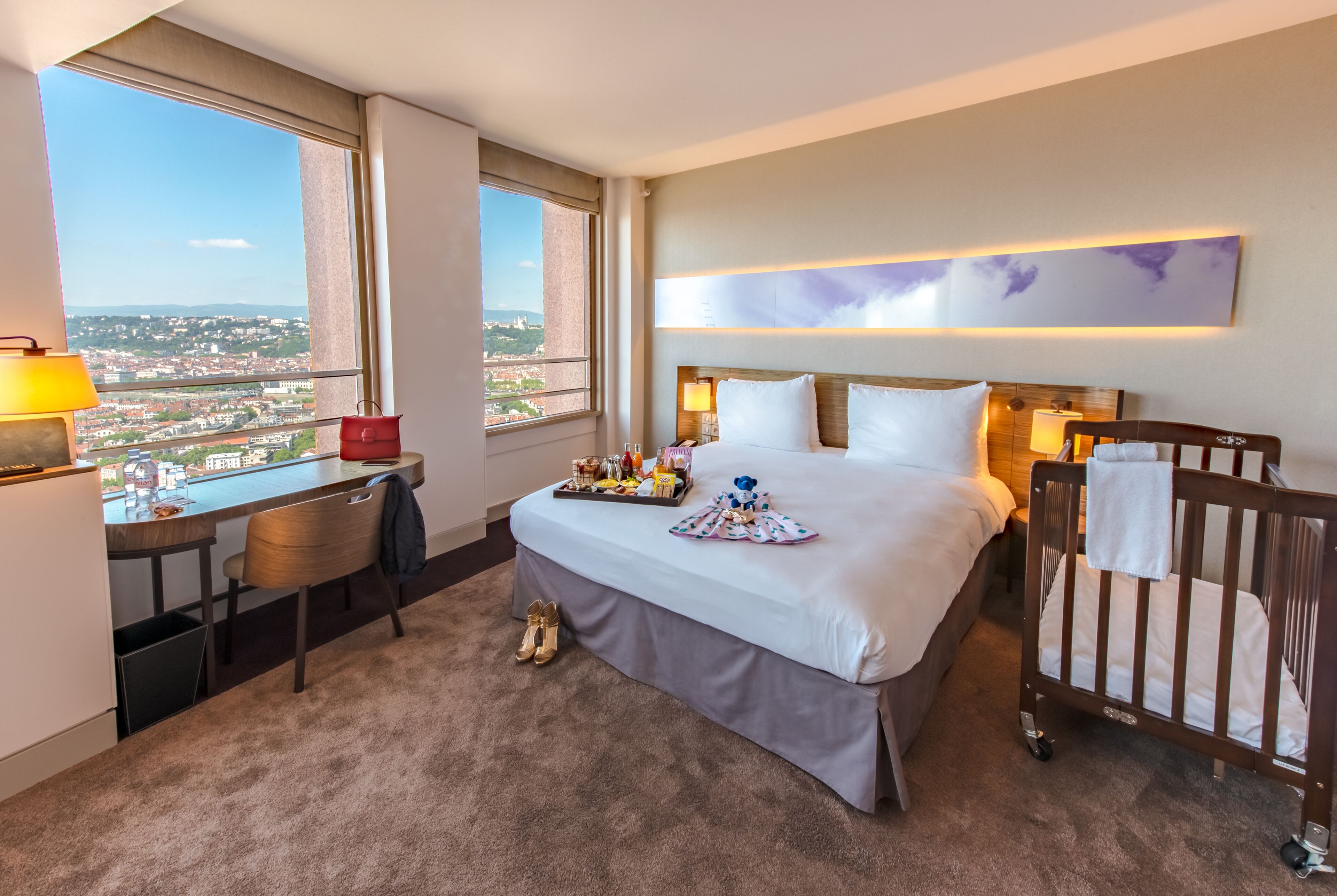 A hotel room with a bed, a desk, a chair, a crib, and a city view.