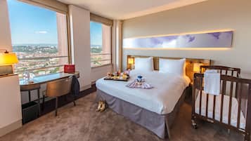 Quarto de hotel com uma cama, escrivaninha, cadeira, berço e vista para a cidade.