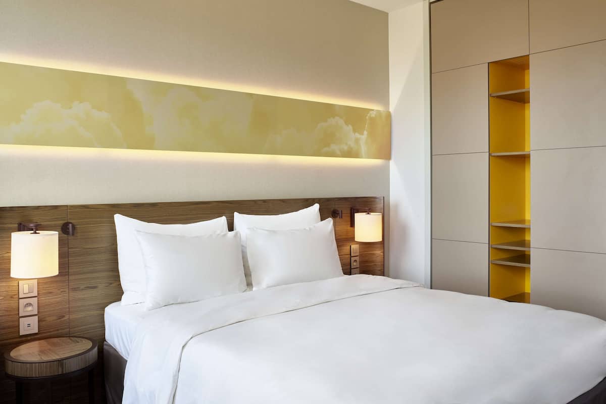 A hotel room with a neatly made bed, two bedside lamps, a round nightstand, and a wall-mounted shelf.