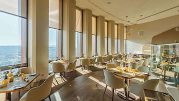 A modern restaurant with high ceilings and large windows, offering a view of the cityscape. Tables are neatly arranged with chairs, and the floor is made of wood.