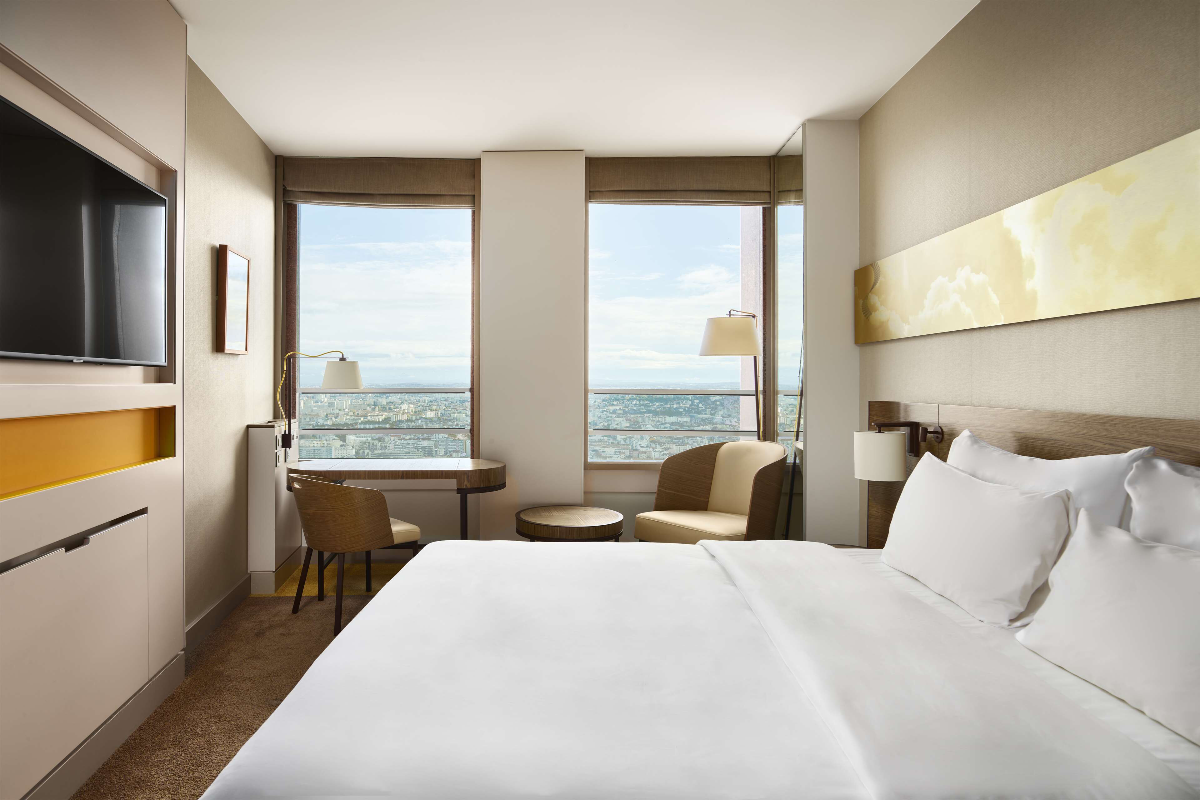 A hotel room with a large bed, a desk, two chairs, a TV, and a city view.