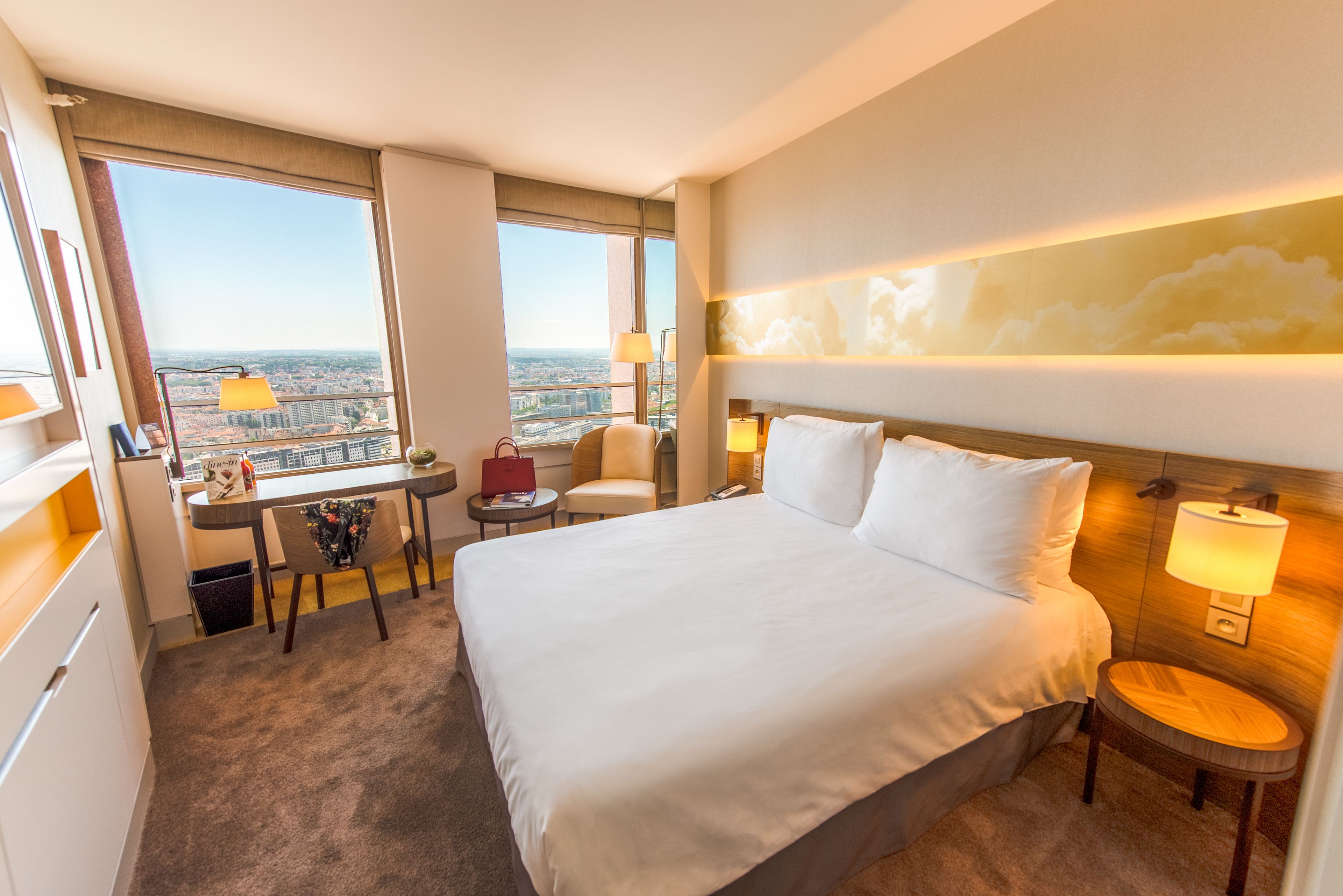 Habitación de hotel con una cama grande, un escritorio, una silla y vistas a la ciudad a través de un amplio ventanal.